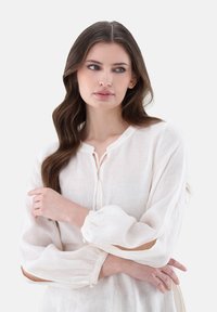 Weiße Leinenbluse mit langen, puffartigen Ärmeln, dekorativen Ausschnitten an den Ellbogen und einem Rundhalsausschnitt mit Bindebändern vorne.