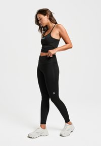 Zwarte sportbeha met dunne bandjes, gecombineerd met zwarte high-waisted legging. Bevat een subtiel logo en een glad, stretchy materiaal. Witte sport schoenen.