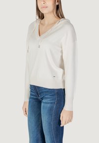 Felpa bianca con cappuccio, scollo a V e maniche lunghe, con orlo a coste e coulisse con dettagli metallici, abbinata a jeans blu.