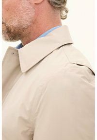 Beige waterproof jacket with a classic collar, featuring a smooth texture, and an understated design. Blue shirt visible underneath.
