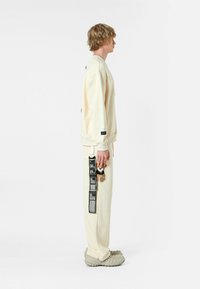 Sudadera crema de mangas largas y corte holgado, combinada con joggers a juego que presentan una etiqueta gráfica lateral y un llavero. Calzado texturizado.