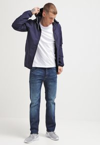 Marineblå vandtæt jakke med hætte, båret over en hvid t-shirt, parret med mørke denim jeans og grå sportssko.