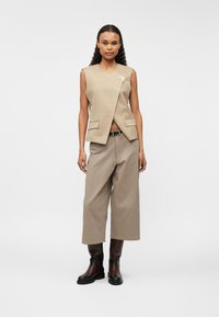 Bēšs bezpiedurkņu veste ar V veida kakla izgriezumu un priekšējām kabatām, apvienota ar platām bēšām biksēm un tumši brūnām ādas potīšu zālēm.