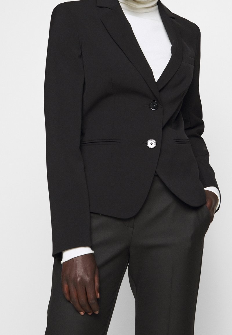 Personne portant un blazer noir taillé sur mesure par-dessus un col roulé blanc avec un pantalon noir, une main dans la poche et l'autre suspendue.
