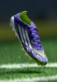 Purple and neon green soccer cleat with "F50" branding, suspended above grass on a marked sports field.