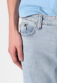 Világoskék farmernadrág, klasszikus öt zsebes dizájnnal, enyhe kopással és eltérő árnyalatú cérnával. Kéz a combon.