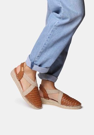 Brown woven espadrille sandals with a beige cross-strap design and jute soles. Light blue rolled denim pants are worn with the shoes.