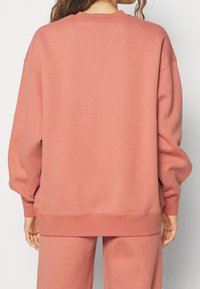 Persona che indossa un set coordinato di felpa e pantaloni in rosa polveroso, mostrata di spalle su uno sfondo neutro.