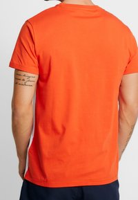 Muž ve svetle oranžovém tričku s krátkým rukávem a tmavých kalhotách, zobrazený z pohledu ze zad se viditelnými tetováními na obou pažích.