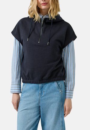 Femme portant un pull sans manches à capuche bleu marine avec fermeture éclair, par-dessus une chemise rayée bleue et un jean bleu clair, sur fond blanc.