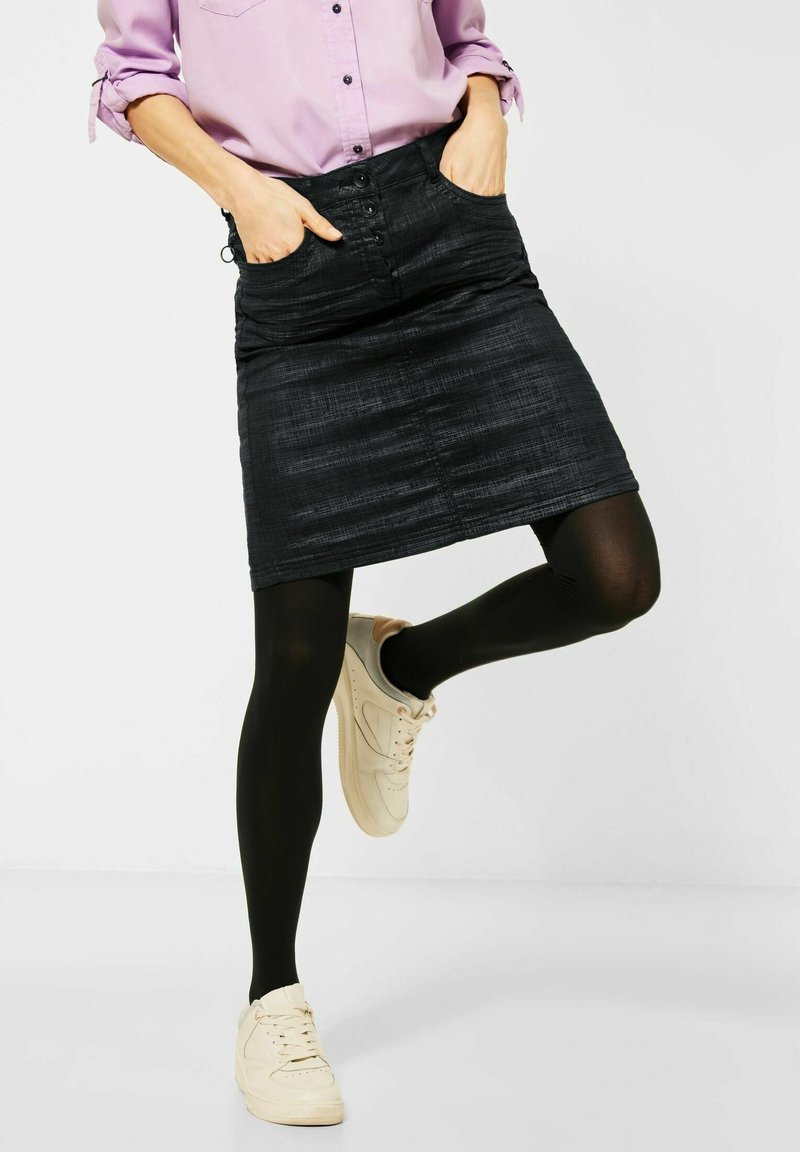 Černá džínová tužková sukně s předním zapínáním na knoflíky, bočními kapsami a texturovaným povrchem, doplněná o černé punčocháče a béžové tenisky.
