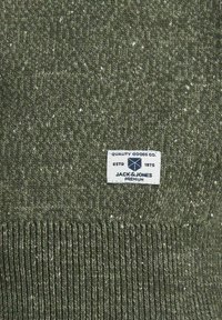 Maglione a collo alto di colore verde oliva, lavorato a maglia con un motivo testurizzato. Include un'etichetta bianca con testo blu che dettagli il marchio e la qualità. Orlo a coste.