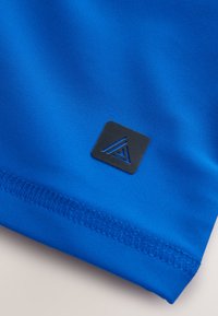 Klart blåt stof med glat tekstur; har en sort logo-patch med geometrisk design, placeret nær kanten med flad syning.