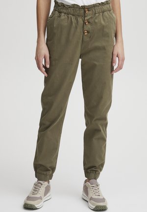 Olivgrön, byxor med elastisk midja och avslappnad passform, med tre träknappar och elastiska muddar. Kombinerade med grå sneakers.