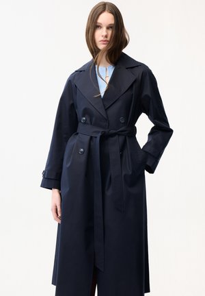 Ung kvinde med langt brunt hår iført en mørkeblå trenchcoat med bælte over en lyseblå top mod en ensfarvet hvid baggrund.