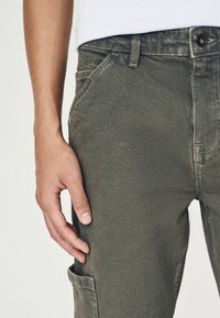 Pantaloni cargo de culoare verde măsliniu, dintr-un material texturat, dotati cu buzunare laterale și cusături vizibile. Închidere cu buton la talie.