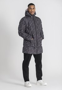 Chaqueta acolchada negra con patrón de logotipos blancos, con un alto collar y cierre de cremallera. El modelo lleva pantalones negros y zapatillas blancas.