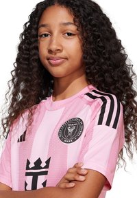 Maillot de football rose avec des rayures noires et un logo, présentant une texture en maille respirante. Le design comporte un col ras du cou et des manches courtes.