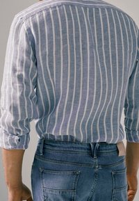 Chemise en lin rayée bleu et blanc avec manches retroussées, associée à un jean en denim bleu avec poches arrière et une étiquette à la taille.