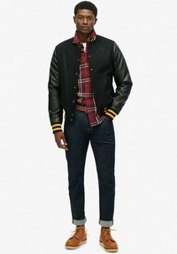 Zwart varsityjack met leren mouwen, gele strepen op de manchetten, een geruit shirt eronder, donkere spijkerbroek en bruine veterschoenen.