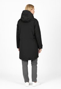 Lang sort pufferjakke med hætte, glat tekstur og let buet kant. Båret med grå bukser og hvide sneakers, set fra ryggen.