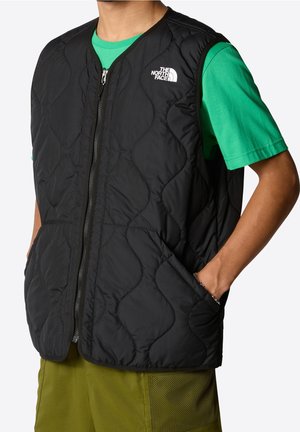 Gilet trapuntato nero con chiusura a zip e tasche, caratterizzato da un logo sul lato sinistro del petto. Indossato sopra una maglietta verde.