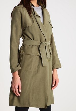 Vrouw draagt een olijfgroene trenchcoat met een ceintuur en klepzakken over een grijze top, staand met handen ontspannen langs het lichaam.
