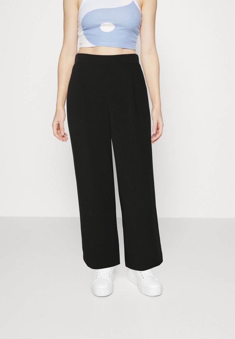 Schwarze weite Hosen aus weichem Stoff, mit vorderen Falten und einem glatten Taillenbund, kombiniert mit einem hellblauen Crop-Top und weißen Turnschuhen.
