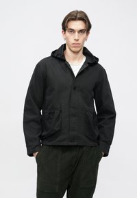 Jachetă neagră cu glugă, închidere cu nasturi, buzunare cu șnur și material texturat. Poartă peste un tricou alb și pantaloni din catifea verde închis.