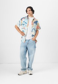 Cămașă cu mâneci scurte, cu un model multicolor, peste un tricou alb, asortată cu blugi largi de culoare albastru deschis și adidași bej. Texturi de țesătură netedă.
