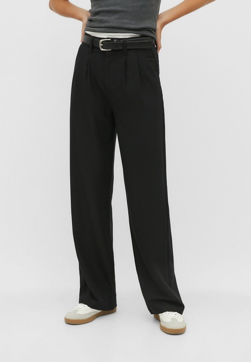Personne portant un pantalon noir à jambes larges avec une ceinture noire, un haut gris sans manches et des baskets blanches, debout les mains sur les hanches.