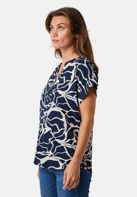 Dunkelblaue Bluse mit abstraktem weißen Muster, kurzen Ärmeln und V-Ausschnitt, aus glattem Stoff, getragen mit blauen Jeans.