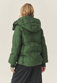 Jachetă puf verde cu glugă, prezentând secțiuni matlasate și manșete ribbate. Designul este robust, cu o textură netedă și colțuri rotunjite.