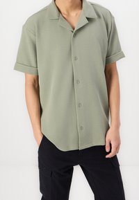 Jasnozielona koszula na krótki rękaw z guzikami i teksturowanym wykończeniem, z klasycznym kołnierzem i zaokrąglonym dołem, noszona z ciemnymi spodniami bojówkami.