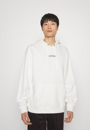 Sudadera blanca de tejido suave con un bolsillo tipo canguro en el frente y el logo "GUESS" en negro en el pecho. Mangas amplias.