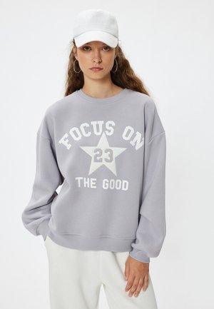 Grå oversized sweatshirt med teksten "FOCUS ON 23 THE GOOD", hvid stjerne grafik, ribbede manchetter og kant. Sammen med en hvid kasket og lyse bukser.