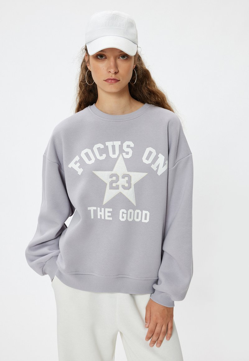 Sudadera oversize gris con el texto "CONCENTRA EN 23 LO BUENO", gráfico de estrella blanca, puños y dobladillo acanalados. Combinada con una gorra blanca y pantalones claros.