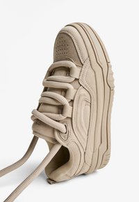 Beige vastag talpú sneaker, párnázott nyakkal, texturált fűzőkkel és varrott panel részletekkel, fehér háttér előtt, ferde nézetben bemutatva.