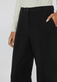 Zwarte maatwerkbroek met een recht model, voorzien van een knoopsluiting, verticale plooien en zijzakken voor extra detail.