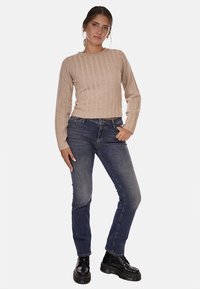 Maglione beige a coste con maniche lunghe, abbinato a jeans blu scuro e stivali neri con lacci. L'outfit presenta uno stile casual.