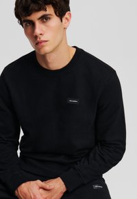 Felpa nera realizzata in morbido tessuto, presenta un classico colletto rotondo e polsini aderenti, con un piccolo patch bianco con logo sul petto.