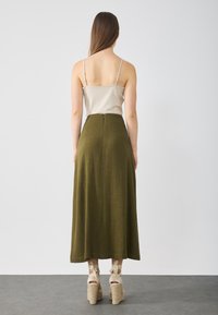 Olive green long skirt with a textured finish, high waist, and a back zipper. Paired with a beige tank top and light-colored wedge heels.
