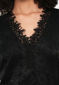 Crna bluza od čipke s V-izrezom i detaljnom čipkastom oblogom, mekom teksturom i cvjetnim uzorcima po cijelom materijalu. Lagani i prozirni dizajn.
