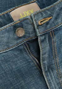 Mėlyni džinsai su dalinai atidarytu užtrauktuku, ruda siuvinėta sagos anga ir kreminės spalvos etiketė su geltonu tekstu „JJXX“.