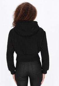 Fekete, rövidített kapucnis pulóver puha anyagból, kapucnival, hosszú ujjakkal és karcsúsított derékrésszel. A hátsó nézet sima textúrát és tiszta vonalakat mutat.