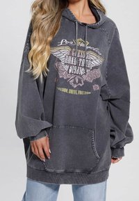 Femme portant un sweat à capuche noir délavé et surdimensionné avec un motif ailé indiquant "Los Angeles, Guess Road Tour 1981" et un jean bleu, les cheveux lâchés sur l'épaule.