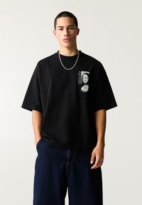 Schwarzes Baumwoll-T-Shirt mit kurzen Ärmeln, das auf der linken Seite ein graues grafisches Design eines Frauengesichts und Text zeigt. Kombiniert mit blauen Jeans.