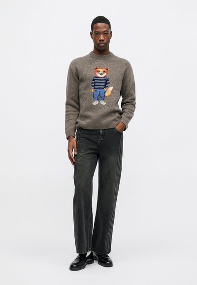 Vīrietis, tērpies pelēkā džemperī ar lapsu svītrainā kreklā un džinsos, melnajās platās biksēs un melnās kurpēs, stāv uz balta fona.