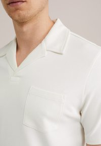 Kortärmad, gräddfärgad pikétröja i mjukt tyg, med spridd krage och en bröstficka med subtila sömnadsdetaljer.