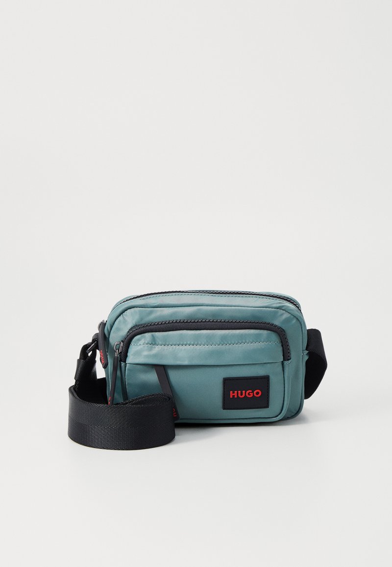 Geantă crossbody texturată de culoare teal cu accente negre, având două compartimente cu fermoar și o curea ajustabilă, patch cu logo roșu.